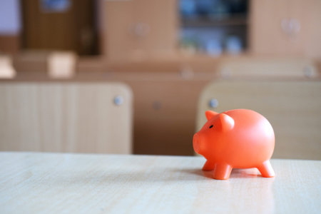 Bright orange piggy bank sits on a wooden table in a school classroom, representing the importance of saving for educational expenses and financial literacyの写真素材