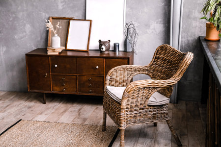 Modern living space with a wicker chair next to a wooden sideboard displaying decorative items and empty frames against a textured gray wallの写真素材