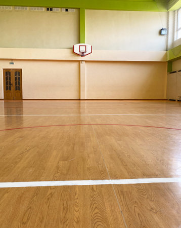 Interior view of a basketball court showcasing a wooden floor with red and white lines, a wall-mounted hoop, and a green ceiling in a gymnasiumの写真素材