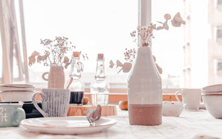 Bohemian style table setting with ceramic dishes, glass bottles, and dried flowers on a light tablecloth, illuminated by natural light from a nearby windowの写真素材