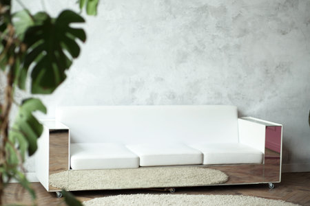 Contemporary white sofa with reflective sides sits on a round rug in a minimalist room, complemented by a leafy plant and a textured wallの写真素材