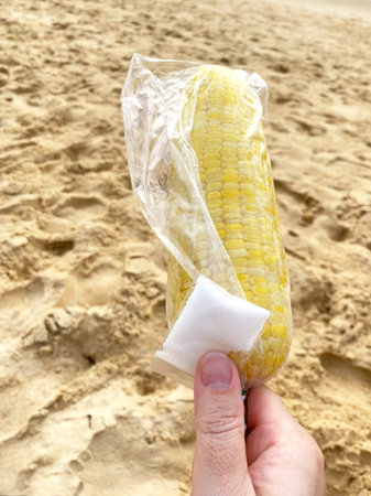 Boiled corn on the beach is held in a hand, wrapped in plastic with a salt packet, sandy ground and ocean waves visible in the backgroundの写真素材