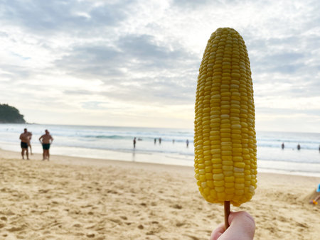 A hand presents a grilled corn on a stick at the beach, with people swimming in the ocean and a...