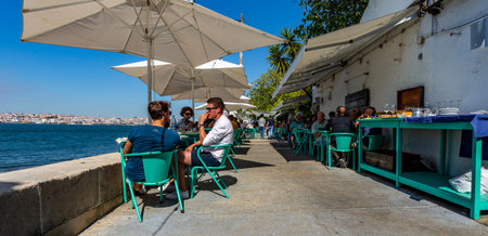 People enjoying the good summer weather at an open-air restaurant by the Tagus River in Almada,...