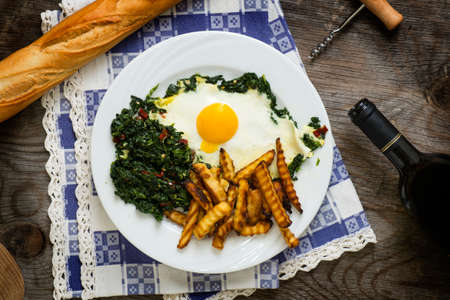 Plate with fried egg, spinach and potatoes placed on a rustic table with bread and wineの写真素材