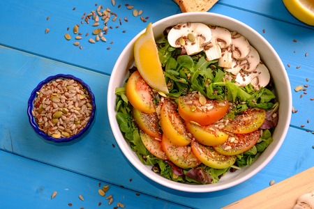 Top view of a bowl of mixed salad with tomatoes, mushrooms, lettuce and seeds with bread on a wooden tableの写真素材