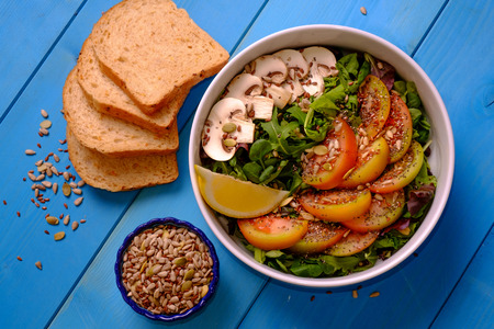 Top view of a bowl of mixed salad with tomatoes, mushrooms, lettuce and seeds with bread on a wooden tableの写真素材