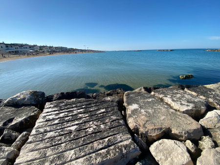 Tranquil seascape showing rocks in the foreground, a beach with people and buildings in the background, under a clear blue sky in Francavilla al Mareの写真素材