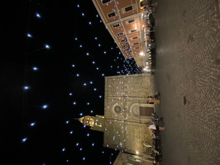 Nighttime illumination highlighting Atri Cathedral's ornate facade and bell tower, casting warm glow over historic Piazza Duomo with visitors lingering beneath festive lightsの写真素材