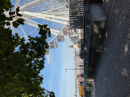 White ferris wheel cabins rotating against a clear blue sky, overlooking the cityscape of Antwerp, with tree branches and cobblestone street in the foregroundの写真素材