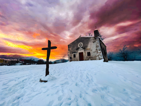 Capturing the beauty of a snowy landscape, featuring a wooden cross and a quaint church, bathed in the warm hues of sunsetの写真素材