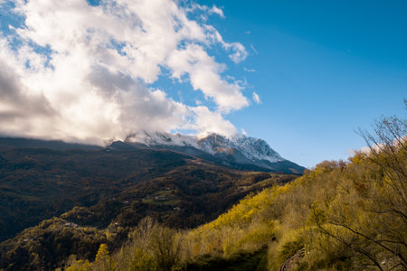 Changing seasons with an autumn landscape featuring a snowy mountain range, vast blue sky, and white clouds above green-yellow hillsの写真素材