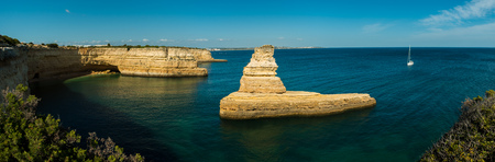 Panorama of the Yellow Submarine Rock on the Algarve Coast of Portugal, in the bay of Praia da Morena, with a sailing boat heading towards it under a clear blue sky.の写真素材