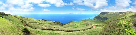 Large panorama overlooking cows grazing next to a winding road in the SÃ£o Jorge countryside with the island of Pico in the background. Pico summit is seen across the ocean under a dramatic cloudy sky.の写真素材