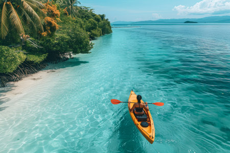 Aerial view of a person kayaking in clear turquoise waters near a lush tropical island coastline with vibrant foliage.の素材