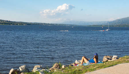 TOURIST SUNBATHING ON THE SHORE OF A LAKE WITH SAILBOATS IN THE BACKGROUNDの写真素材