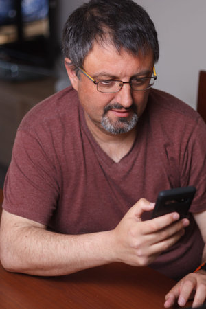 Gray-haired man with glasses and goatee is looking intently at his mobile phone wearing a red...