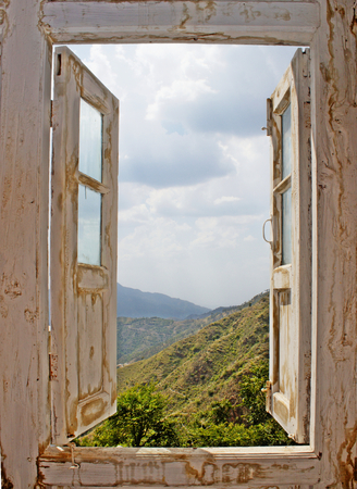 View of the mountains and clouded sky from an open old white wood window. Ideal for concepts related to outdoors, nature, exploration, peace, and inspiration.の写真素材