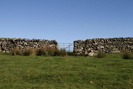 old metal gate in a dry stone wall in a green grass field and blue sky.の写真素材