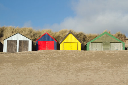 Multi-coloured beach huts with sand banked up against the doors, the scene is backed by a dune with blue sky and cloud.の写真素材