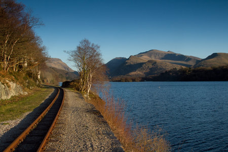 A single track rail line next to a lake with trees and mountains in the distance with a blue sky.