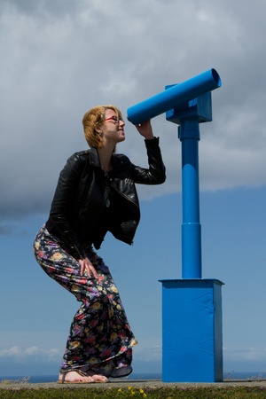 A beautiful woman in floral pants and black leather leather jacket looks through a blue telescope.の写真素材