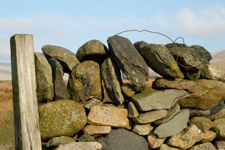 A dry stone wall construction with layers of rock and a coping with rusty wire against a wooden...