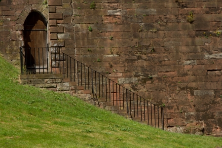 An historic red sandstone castle wall with a set of stairs leading to an arched doorway with mown green grass in the foreground.のeditorial素材