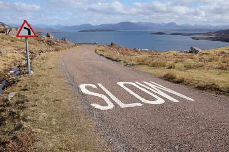 A narrow road with the word 'SLOW' painted on the floor and a triangular sign indicating a bend ahead with the sea and islands in the distance.の写真素材