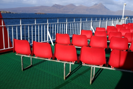 Rows of red plastic seating on a green rubber floor with white railings and a view of the sea and mountains in the distance.の写真素材