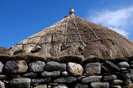 A stone wall with a thatched roof and ropes arranged with stone weights against a blue sky.の写真素材