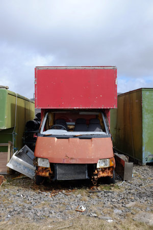 A red van broken and wrecked with a broken windscreen between two green containers against a grey...