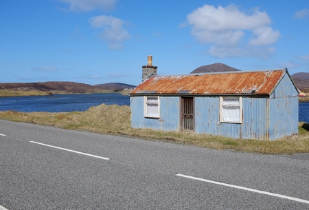 A derelict corrugated metal building on the side of the road with water and hills in the distance under a blue sky.の写真素材