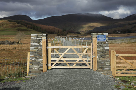 A wooden gate leads to a causeway with a hardcore path flanked by metal fencing with cloud covered...