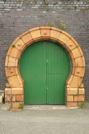 A green painted wooden door with a yellow arch designed with cast brick against an old blue brick wall with plants.の写真素材