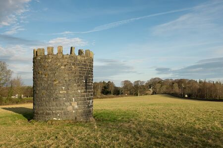 A stone built tower in a grass field leading to trees against a blue cloud sky.の写真素材