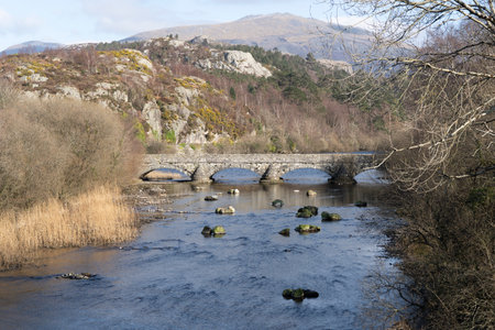 Old bridge over the river in winter time with Padarn Lake and Snowdonia mountains, Wales, UK, in the distance.の写真素材
