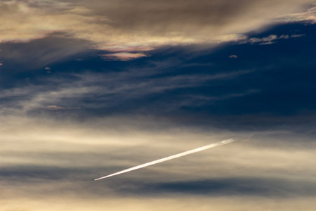 Airplane trail in the blue sky with clouds and sun rays.