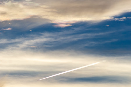 Airplane trail in the blue sky with clouds and sun rays.