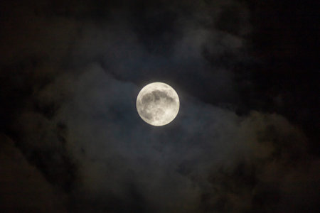 Full moon in the night sky with clouds. The moon is visible through the clouds.