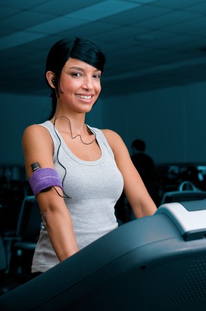 Beautiful young latin woman smiling and listening music while walking on a treadmill at gymの写真素材