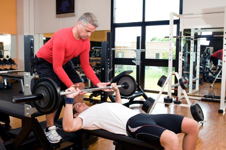 Young man doing weigths on a bench press with the help of the mature instructor at gymの写真素材