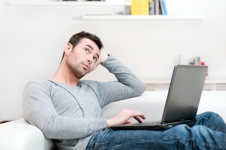 Pensive young man looking up while working on laptop at homeの写真素材