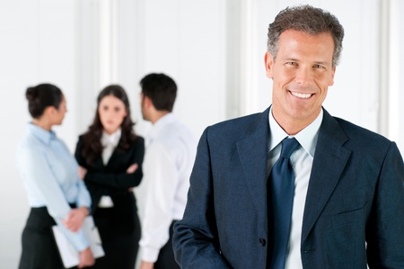Modern mature businessman smiling and looking at camera with his colleagues in the background at officeの写真素材