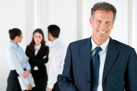 Modern mature businessman smiling and looking at camera with his colleagues in the background at office