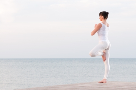 Young healthy woman standing on one leg while meditating and praying outdoor at seaの写真素材