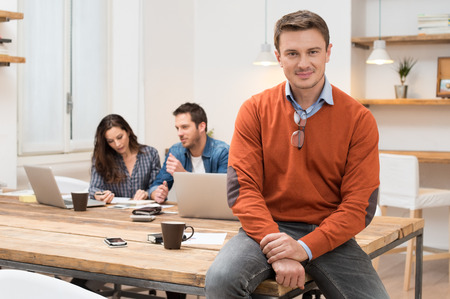 Successful businessman smiling and looking at camera with other workers sitting in backgroundの写真素材