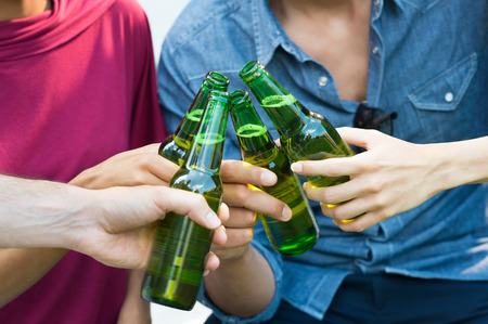 Closeup shot of happy friends toasting with bottles of beer. Girls and guys drinking beer outdoor in a summer day. Close up of female and male hand holding a bottles of beer.の写真素材