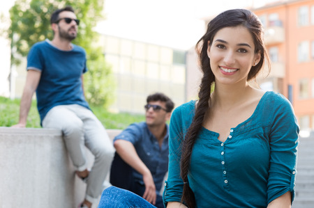 Closeup shot of young woman looking at camera. Portrait of smiling girl in casual looking at camera with her friends in background. Shallow depth of field with focus on beautiful young woman smiling.の写真素材
