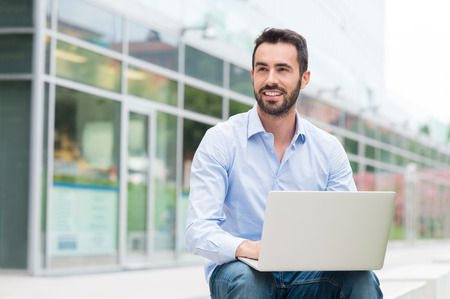 Portrait of young man sitting outside with laptop. He's looking away. Shallow depth of field with focus on young man sitting with laptop.の写真素材